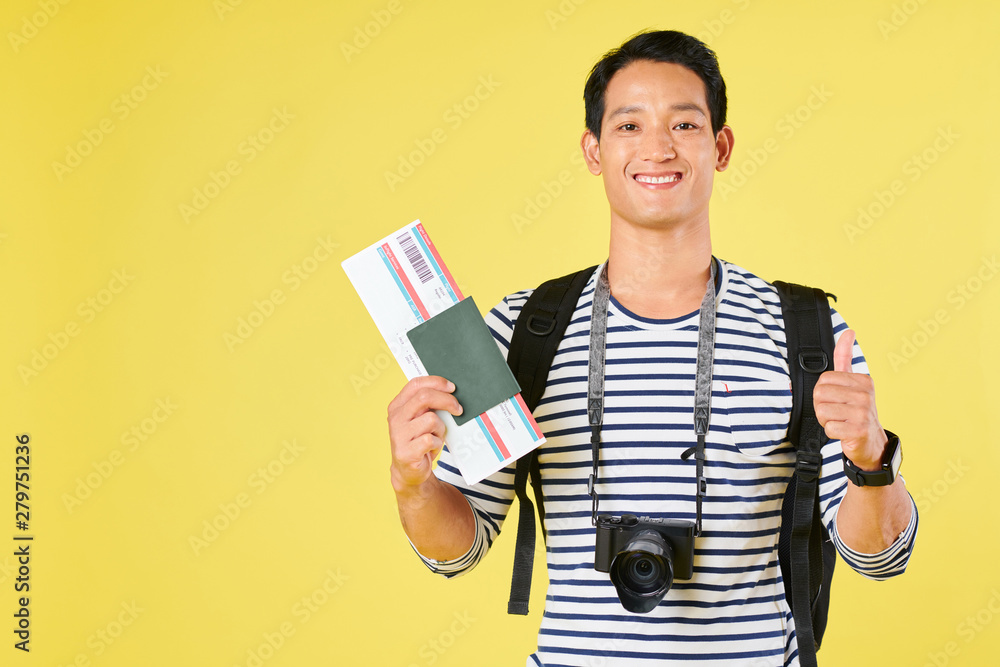 Portrait of positive young Asian tourist with digital camera showing passport and plane ticket, isolated on yellow Portrait of positive young Asian tourist with digital camera showing passport and plane ticket, isolated on yellow