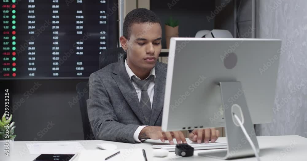 Young african man in a modern office, working at the computer, screens with financial infographics on background, manager at work. Young african man in a modern office, working at the computer, screens with financial infographics on background, manager at work.