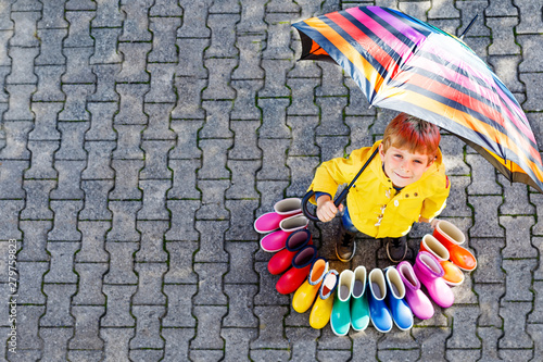 Little kid boy and group of colorful rain boots. Blond child standing under umbrella. Close-up of schoolkid and different rubber boots from high angle. Footwear for rainy fall