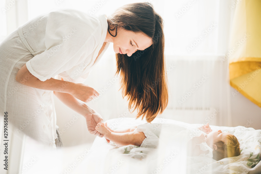 Adorable two month old baby girl lying on the bad. Mother holding daughter legs and smiling. Mother standing near the bed. Concept photo parenthood and motherhood. - obrazy, fototapety, plakaty 