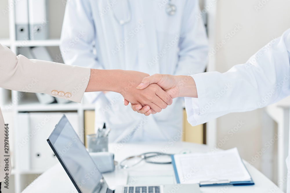 Doctor shaking hand of female patient over office table with opened laptop, selective focus Doctor shaking hand of female patient over office table with opened laptop, selective focus