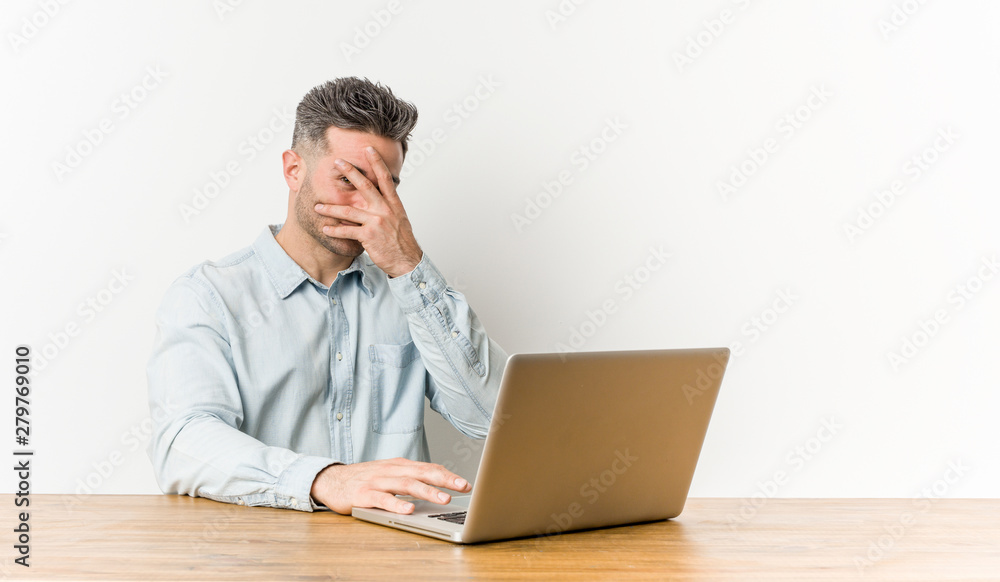 Young handsome man working with his laptop blink at the camera through fingers, embarrassed covering face. Young handsome man working with his laptop blink at the camera through fingers, embarrassed covering face.