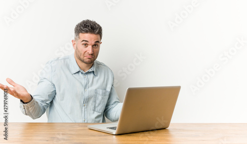 Young handsome man working with his laptop doubting and shrugging shoulders in questioning gesture.