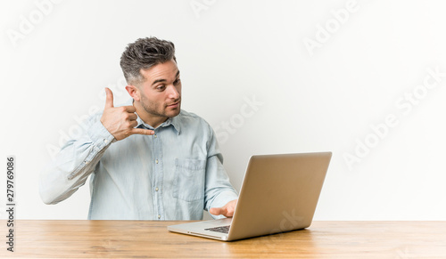 Young handsome man working with his laptop showing a mobile phone call gesture with fingers.