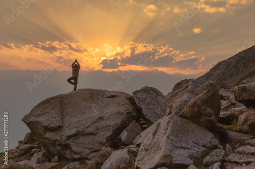 A man performs Yoga (vrikshasana - Tree pose) on a rocky mountain cliff at Deo Tibba trek in Manali, Himachal Pradesh. Serene view of sunset, clouds & sun rays. Summer Hiking in Himalayas. 