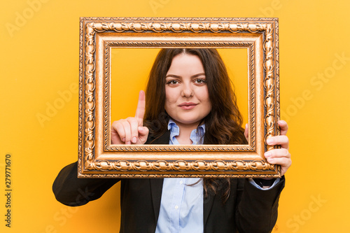 Young curvy plus size business woman holding a frame showing number one with finger. © Asier