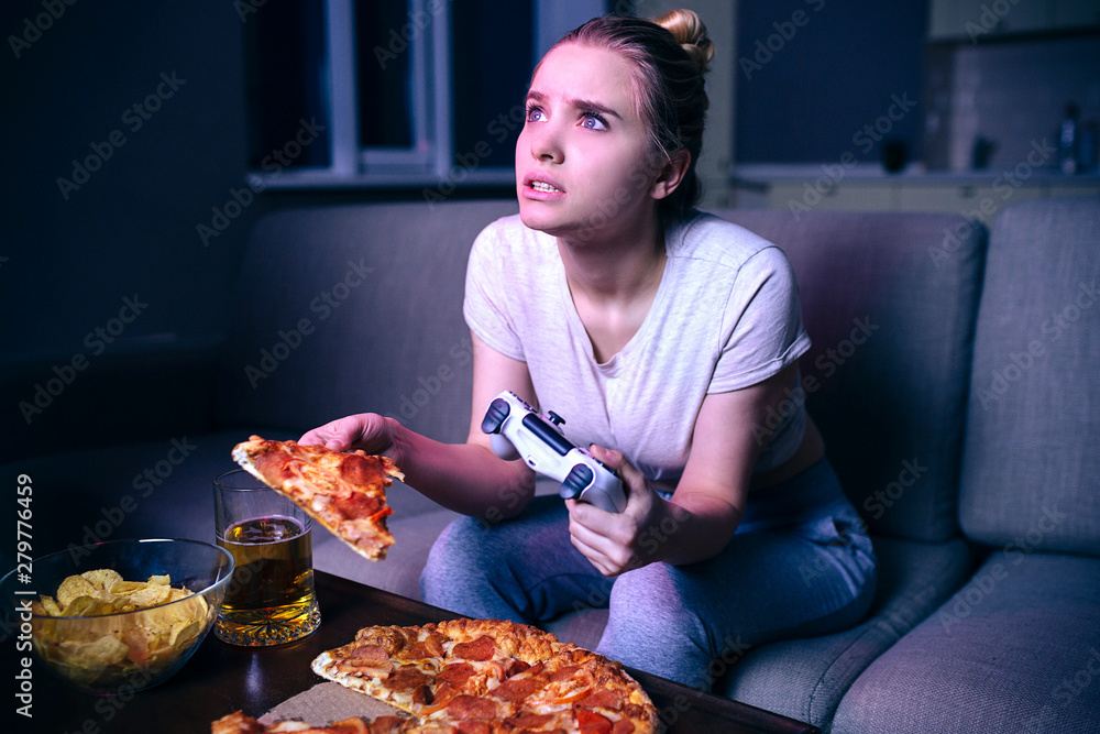 Young woman playing game at night. Hungry model look up on screen. Holding slice of pizza and gamepad. Alone in dark room. Junk food on table.