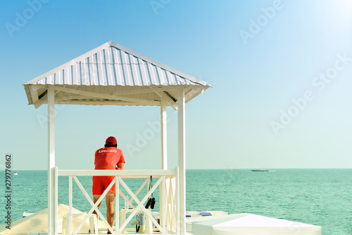 lifeguard on sea beach in watch tower on rescue duty against blue ocean water background back view of person in red t-shirt and swimming shorts with lifeguard label guarding sea shore outdoor activity