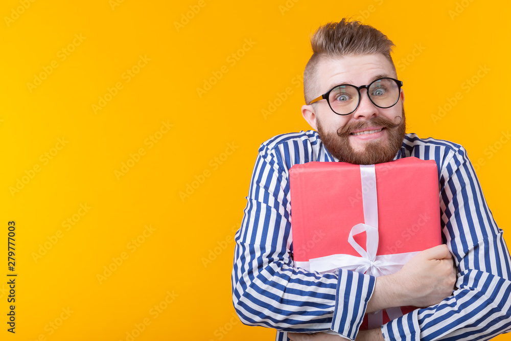 Joyful young hipster guy with a mustache in glasses hugs a red gift box on a yellow background with copy space. The concept of the joy of gifts. Joyful young hipster guy with a mustache in glasses hugs a red gift box on a yellow background with copy space. The concept of the joy of gifts.