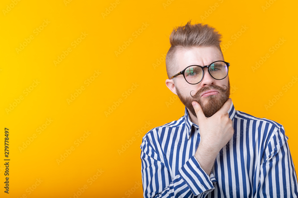 Cute intelligent young man with a beard and with glasses poses on a yellow background with copy space and thinks about something. The concept of a difficult choice.