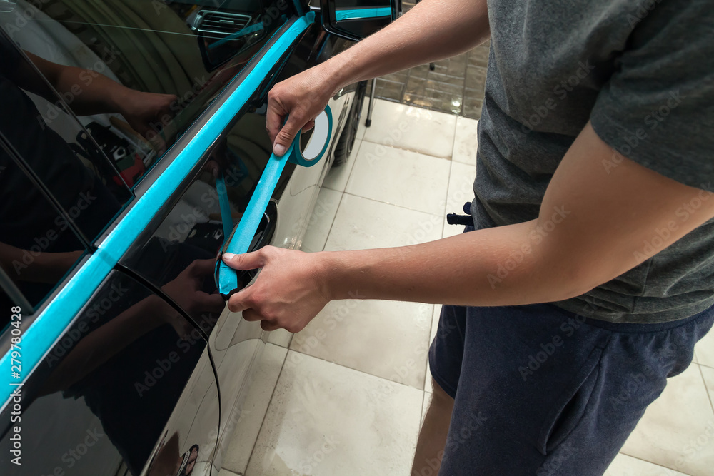 A man of Caucasian appearance with blue paper tape plasters a body element door handle with a black car to polish a paint and varnish and remove scratches in a vehicle wash and detailing workshop. A man of Caucasian appearance with blue paper tape plasters a body element door handle with a black car to polish a paint and varnish and remove scratches in a vehicle wash and detailing workshop.