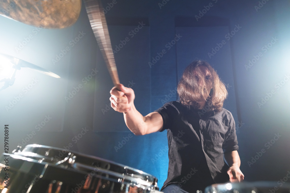 Fototapeta premium Portrait of a long-haired drummer with chopsticks in his hands sitting behind a drum set. Low key. Concepts of the creative freedom of the millenial generation