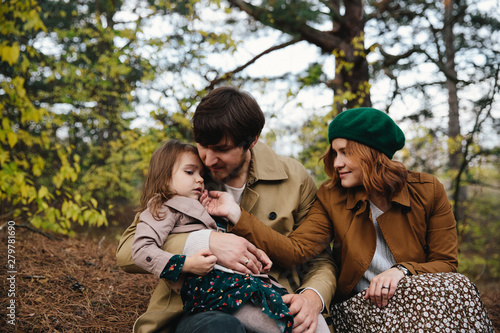 Young father, mother and little toddler daughter girl in a beret and a coat sitting on the ground in the autumn forest. photo