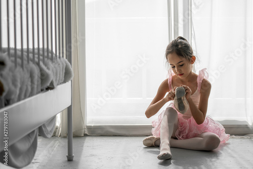 Cute beautiful young little girl ballerina dancer in pink leotard and tutu ballet dress costume sitting on the floor in bedroom wearing and tie ballet slipper shoe for private ballet dance practice.