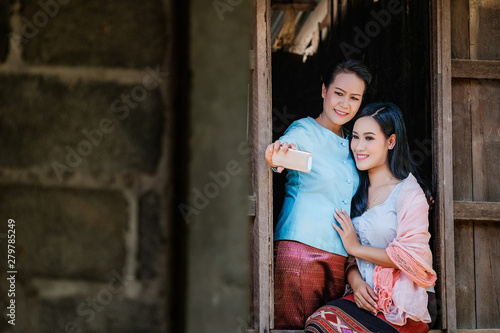 Two Mother and daughter in a traditional Thai dress are taking pictures of themselves with a mobile phone from a wooden window. © subinpumsom