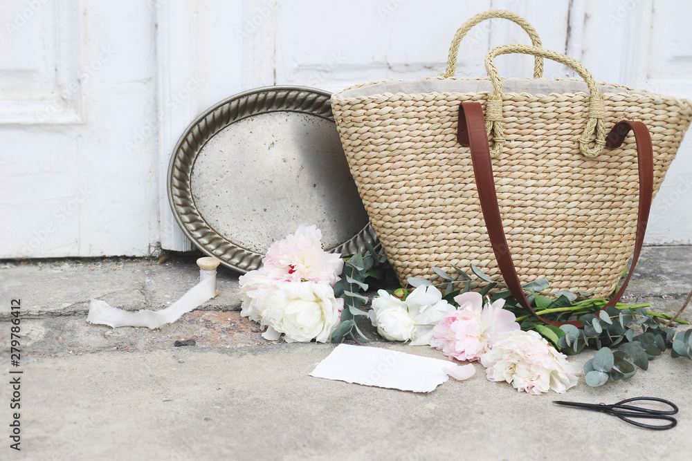 Feminine wedding still life composition with straw French basket bag, pink  peonies flowers, eucalyptus, vintage silver tray with old scissors and silk  ribbons. Old doors and grunge concrete floor. Stock Photo