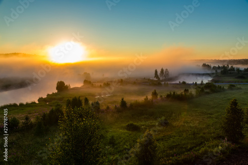Orange sunny light with rays and shadows over the foggy river at dawn. The fog rises above water and stretches to the rising sun. Sunlit misty forest and river in nature