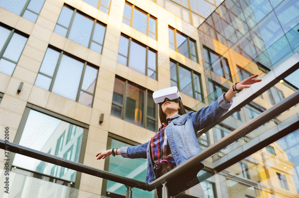 Beautiful girl in denim jacket with long hair playing VR game standing in front of glass building Beautiful girl in denim jacket with long hair playing VR game standing in front of glass building