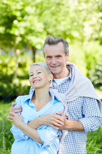 Waist up portrait of happy senior couple embracing and looking at camera while enjoying walk in Summer park