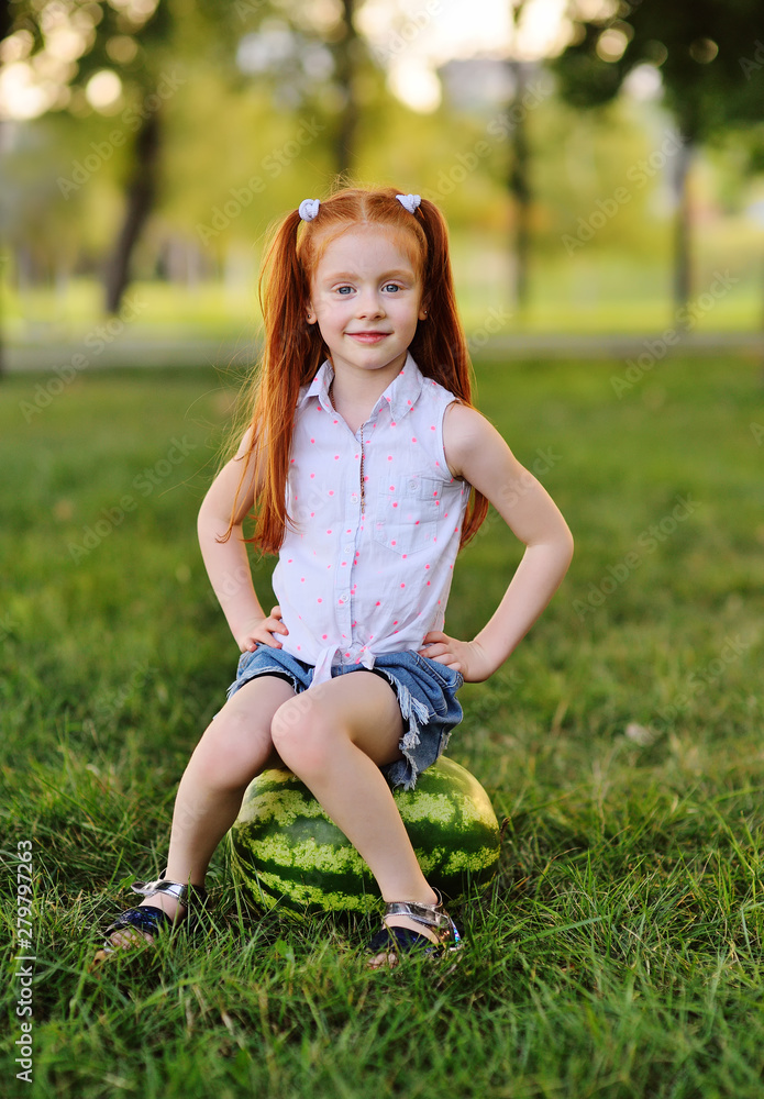 Φωτογραφία baby little funny girl with red hair leaning on a huge watermelon in the Park on