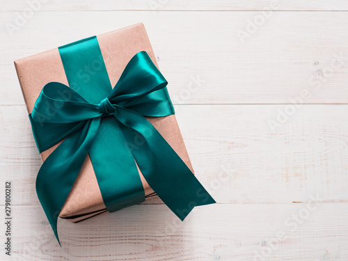 Gift box in craft wrapping paper and green satin ribbon on white wooden table, copy space right. Beautiful Christmas, New Year or Birthday present, flat lay or top view
