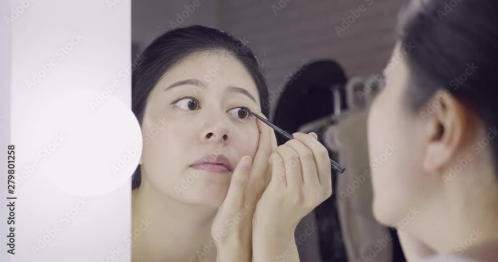 beautiful young dark haired asian woman entertainer using black eyeliner pen while applying makeup on face. close up reflection of girl model from dressing table mirror in backstage room before work beautiful young dark haired asian woman entertainer using black eyeliner pen while applying makeup on face. close up reflection of girl model from dressing table mirror in backstage room before work