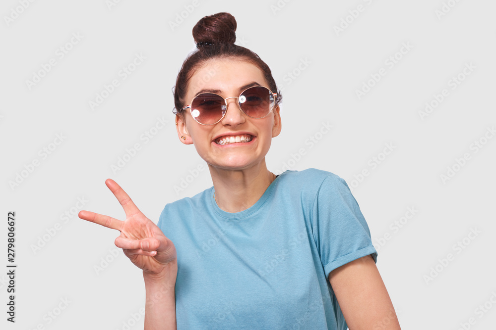 Portrait of happy beautiful girl with toothy smile, wearing blue t-shirt and round sunglasses, posing white studio background. Young student lady standing over grey wall and making peace gesture. Portrait of happy beautiful girl with toothy smile, wearing blue t-shirt and round sunglasses, posing white studio background. Young student lady standing over grey wall and making peace gesture.