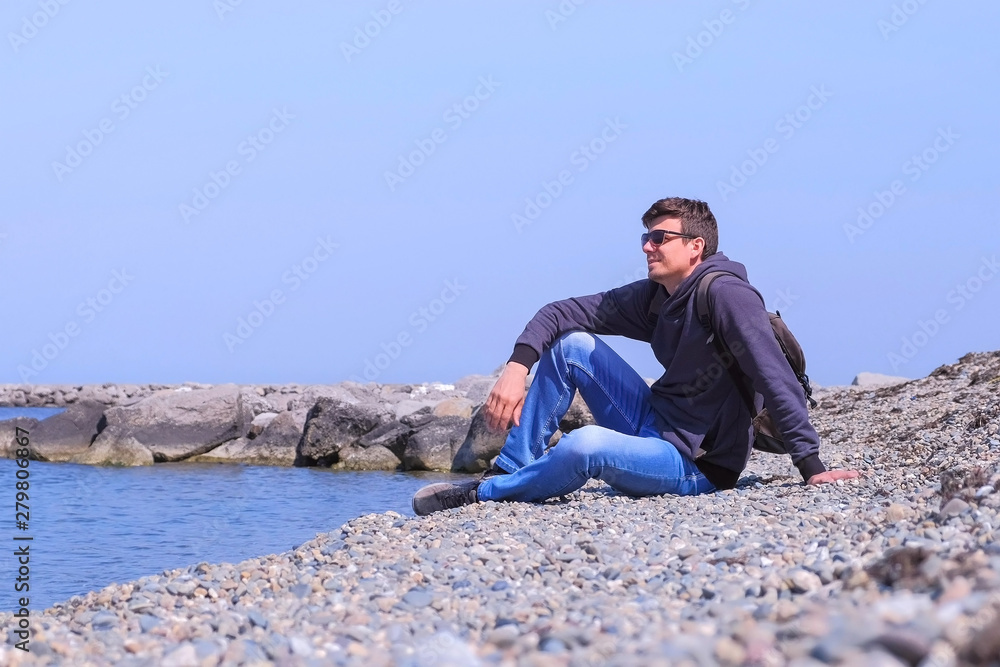 Guy on spring vacation. Happy man tourist traveller sits at stone beach and looks at sea on vacation. He is wearing sunglasses, jeans and sweatshirt. Sunny warm day at seashore. Travel journey trip.