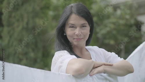 Beautiful mature woman with long hair hanging white clothes on a clothesline looking at camera outdoors. Washday. Lady doing laundry. Concept of sustainabilit. photo