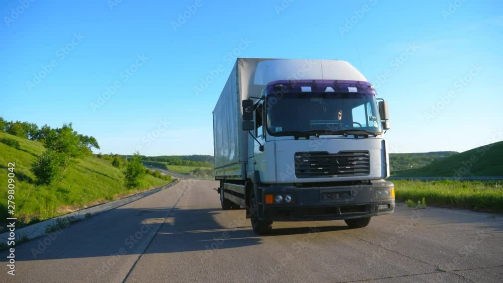 Front view of truck with cargo trailer speeding on highway and transporting goods at sunny day. White lorry riding through countryside road with beautiful nature landscape at background. Slow motion Front view of truck with cargo trailer speeding on highway and transporting goods at sunny day. White lorry riding through countryside road with beautiful nature landscape at background. Slow motion