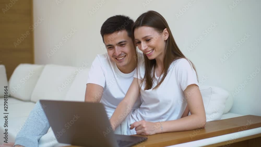 Smiling and cute young couple using laptop in couch at bright living room, online shopping. Happy couple use notebook sitting on white sofa at home Smiling and cute young couple using laptop in couch at bright living room, online shopping. Happy couple use notebook sitting on white sofa at home