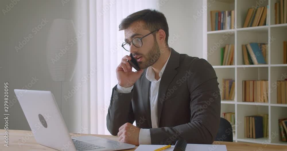 Closeup portrait of adult handsome bearded caucasian businessman in glasses using the laptop and having a phone call in the office indoors Closeup portrait of adult handsome bearded caucasian businessman in glasses using the laptop and having a phone call in the office indoors