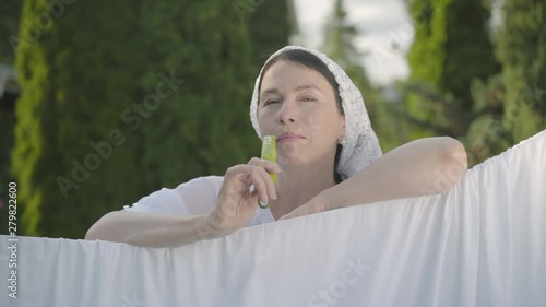 Attractive senior woman with shawl on her head eating green pepper looking at camera smiling over the clothesline outdoors. Washday. Pretty housewife doing laundry. Slow motion. photo