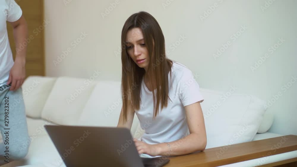 Girl typing on laptop. Guy brings cup of coffee. Young woman drinks coffee. Cute man and female typing on keyboard together sitting on sofa in modern white living room Girl typing on laptop. Guy brings cup of coffee. Young woman drinks coffee. Cute man and female typing on keyboard together sitting on sofa in modern white living room