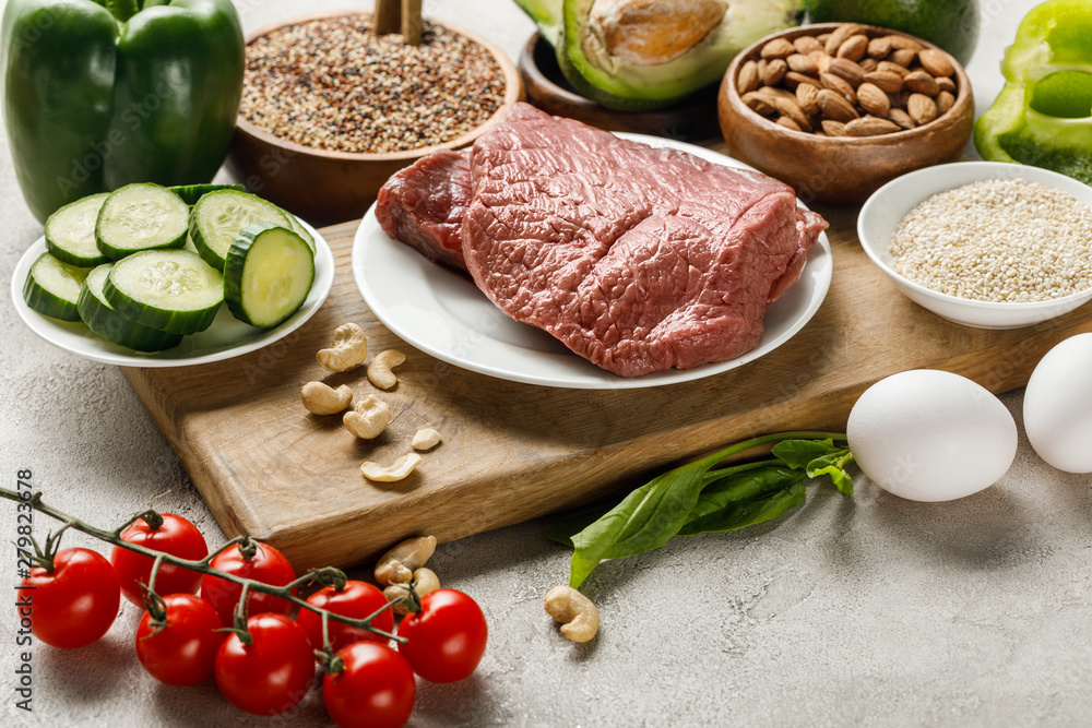 fresh raw meat on wooden chopping board near nuts, vegetables and groats, ketogenic diet menu