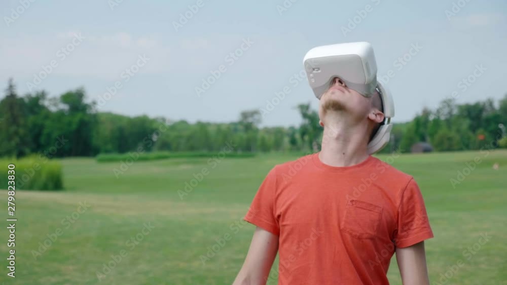A man using a VR headset in the park, dodging obstacles and looking around. A young guy in a red T-shirt and a helmet is experiencing virtual reality against the background of nature. A man using a VR headset in the park, dodging obstacles and looking around. A young guy in a red T-shirt and a helmet is experiencing virtual reality against the background of nature.
