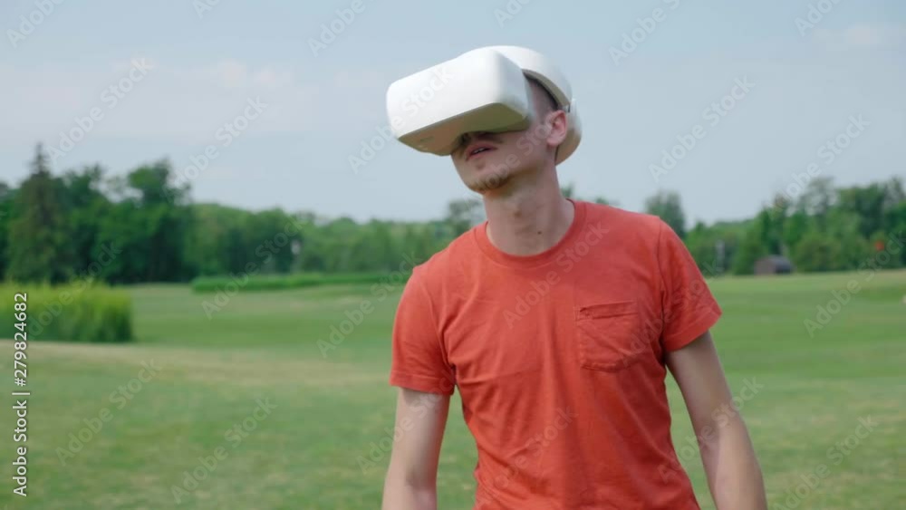A man using a VR headset in the park and looking around. A young guy in a red T-shirt and a helmet is experiencing virtual reality against the background of nature. A human on the right in the frame A man using a VR headset in the park and looking around. A young guy in a red T-shirt and a helmet is experiencing virtual reality against the background of nature. A human on the right in the frame