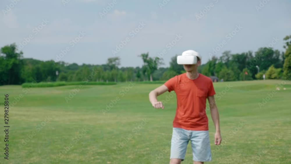 A man using a VR headset and playing a virtual game in the park. Young guy in helmet experiencing augmented reality on the background of nature. A human on the right in the frame. 4K footage. A man using a VR headset and playing a virtual game in the park. Young guy in helmet experiencing augmented reality on the background of nature. A human on the right in the frame. 4K footage.
