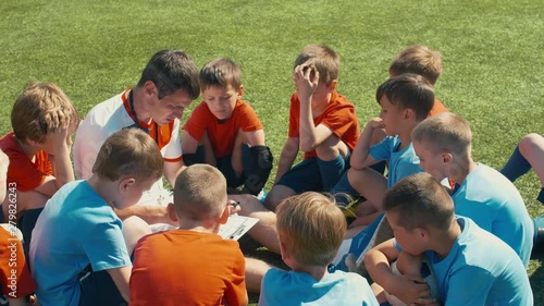 Over-the-shoulder shot of youth soccer team sitting circle on grass around trainer and listening to theory