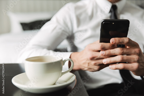 Cropped image of successful man holding smartphone and drinking coffee in hotel apartment during business trip