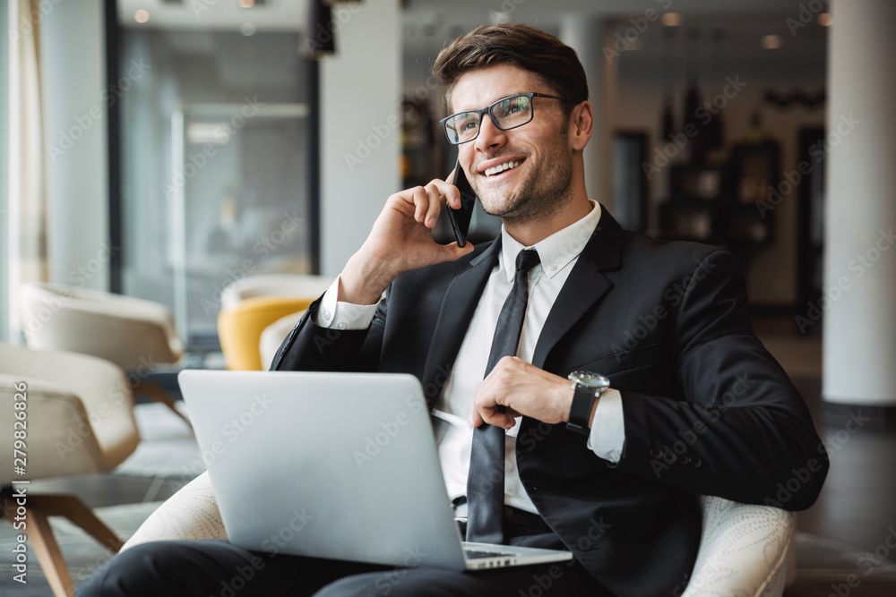 Portrait of happy young businessman using laptop computer and talking on smartphone in hotel hall Portrait of happy young businessman using laptop computer and talking on smartphone in hotel hall