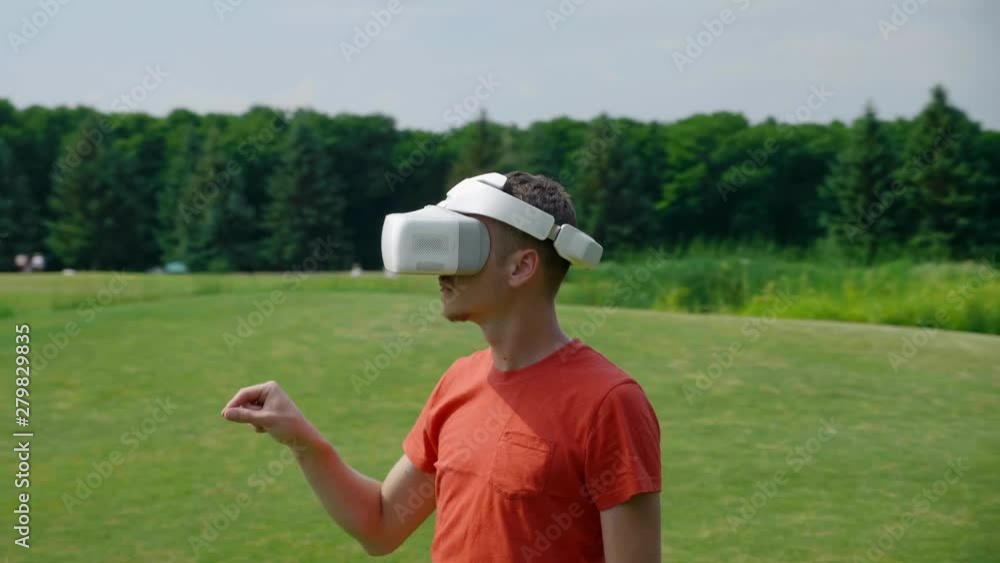 A man using a VR headset and playing a virtual game in the park. Young guy in helmet experiencing augmented reality on the background of nature. Dynamic shooting. 4K footage. A man using a VR headset and playing a virtual game in the park. Young guy in helmet experiencing augmented reality on the background of nature. Dynamic shooting. 4K footage.