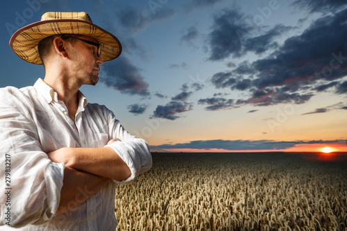 Farmer in a straw hat and glasses standing in a ripe wheat field before sunset. © Volodymyr Herasymov
