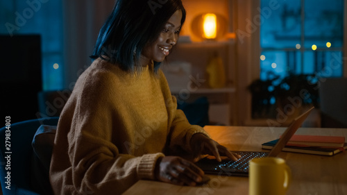 Portrait of Beautiful Black Girl Uses Computer while Sitting at Her Desk at Home  She s Wearing Warm Sweater. In the Evening Creative Woman Works on a Computer In Her Cozy Living Room. 