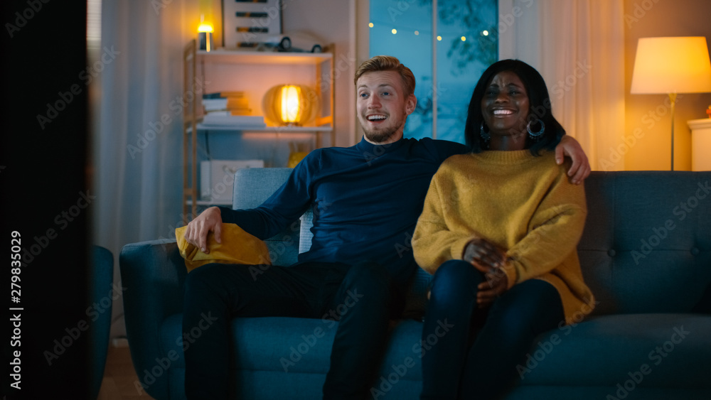 Happy Diverse Young Couple Watching Comedy on TV while Sitting on a Couch, they Laugh and Enjoy Show. Handsome Caucasian Boy and Black Girl in Love Spending Time Together in the Cozy Apartment.
