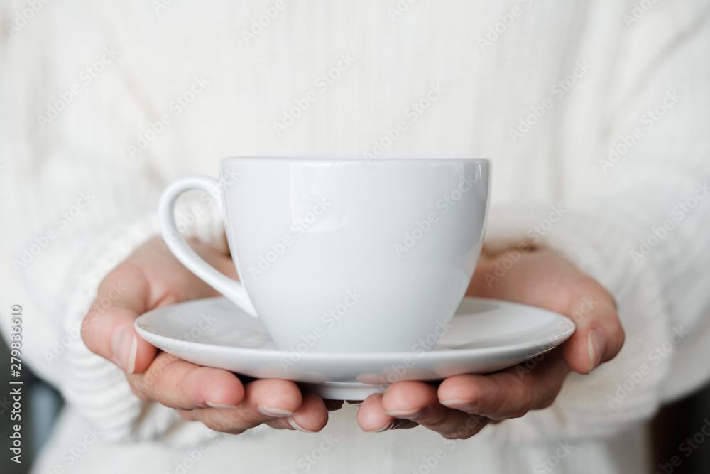 Fototapeta premium Woman in white knitted sweater holding cup of coffee. Female hands with cup of tea. Close up