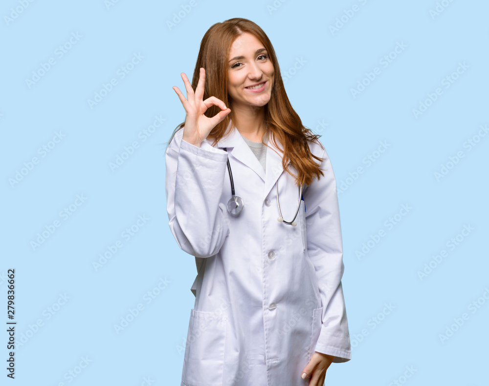 Redhead doctor woman showing an ok sign with fingers on isolated blue background