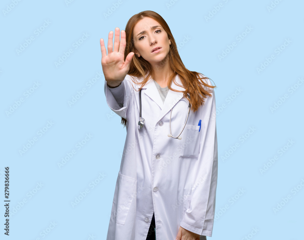 Redhead doctor woman making stop gesture denying a situation that thinks wrong on isolated blue background Redhead doctor woman making stop gesture denying a situation that thinks wrong on isolated blue background