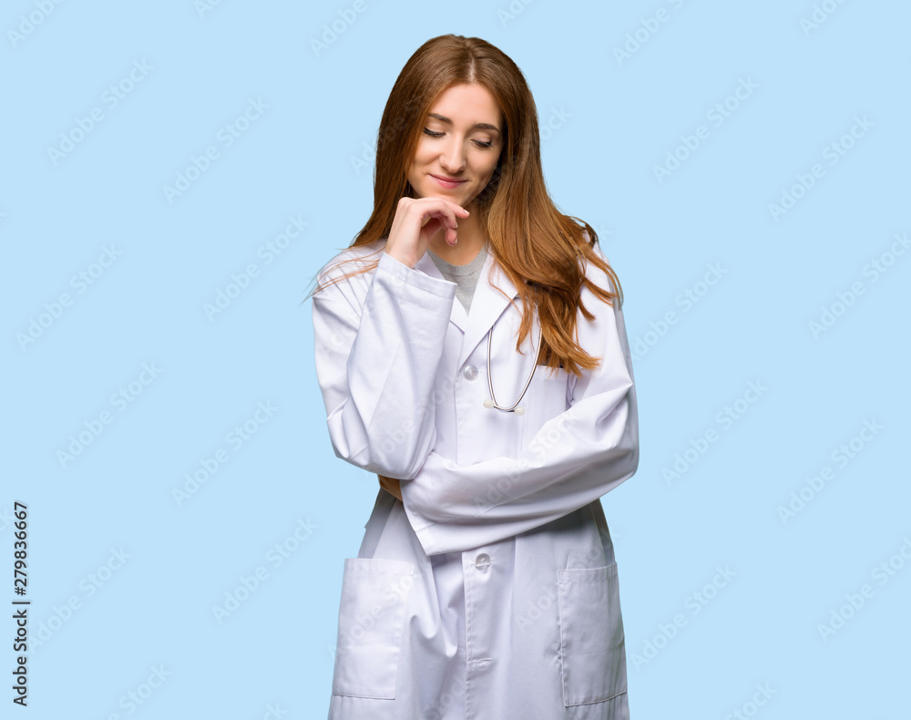 Redhead doctor woman looking down with the hand on the chin on isolated blue background Redhead doctor woman looking down with the hand on the chin on isolated blue background