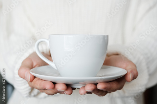 Woman in white knitted sweater holding cup of coffee. Female hands with cup of tea. Close up